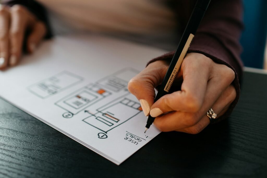 Χέρια που σκιαγραφούν ένα wireframe ιστοσελίδας σε χαρτί δίπλα σε ένα φορητό υπολογιστή